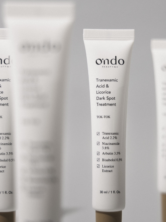 Several tubes of Ondo Beauty's Tranexamic Acid & Licorice Dark Spot Treatment against a light grey background. Some tubes are blurry.