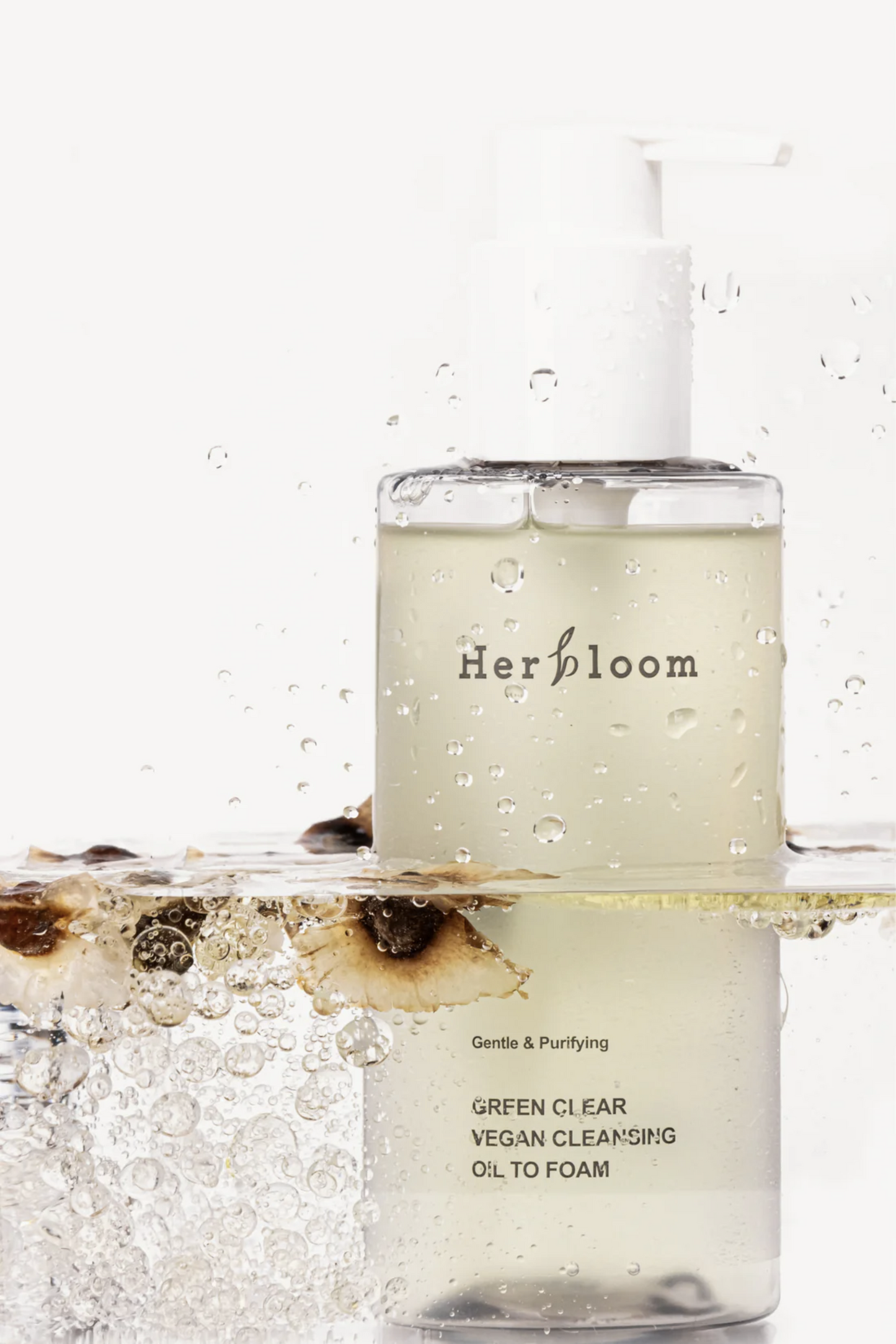 Bottle of Herbloom's Green Clear Vegan Cleansing Oil to Foam submerged halfway in clear water. Light beige petals are floating on the surface. White background. Available at Sinless Skincare.