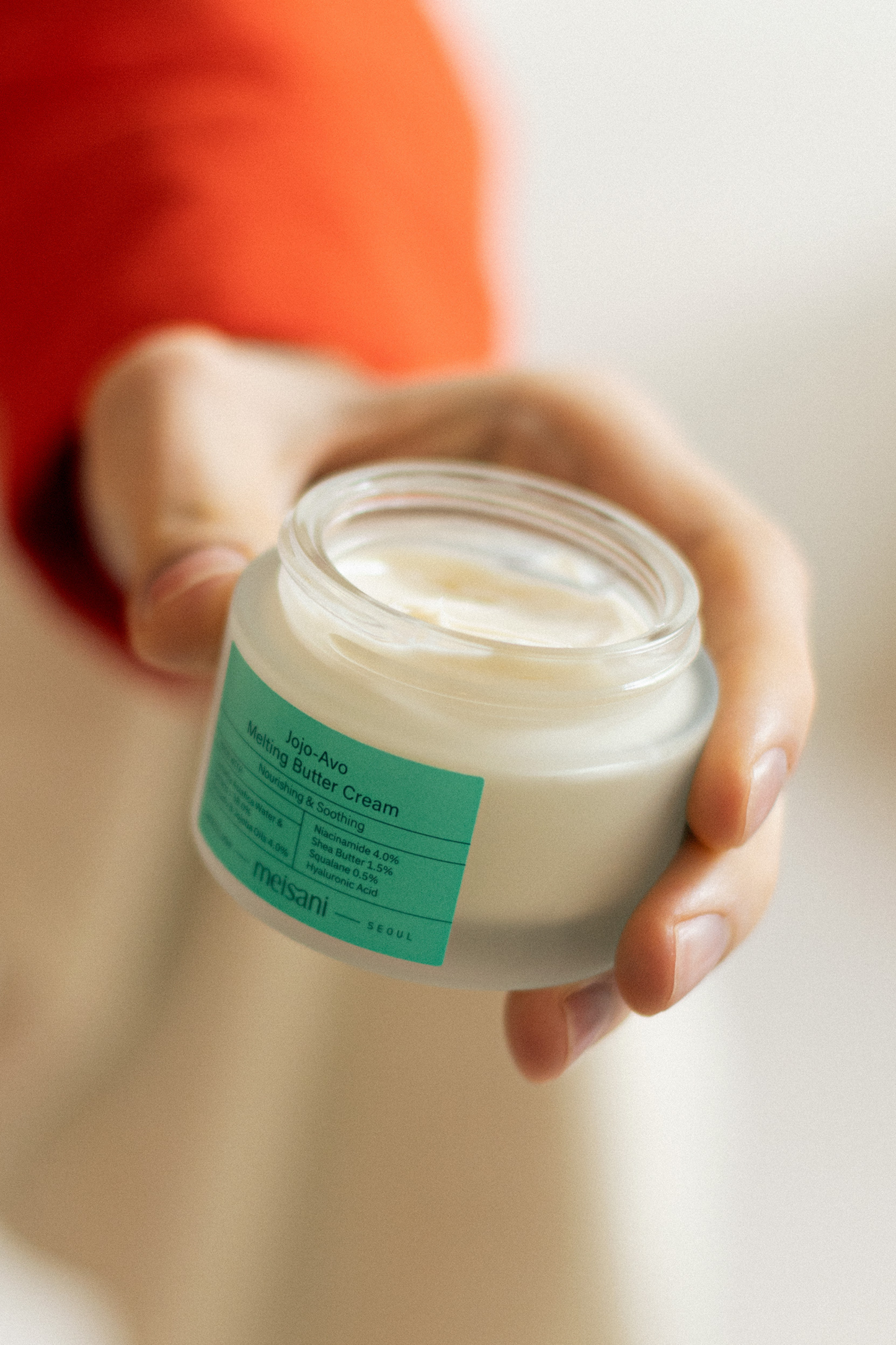 An open jar of Meisani's Jojo-Avo Melting Butter Cream in someone's hand with a blurred background.