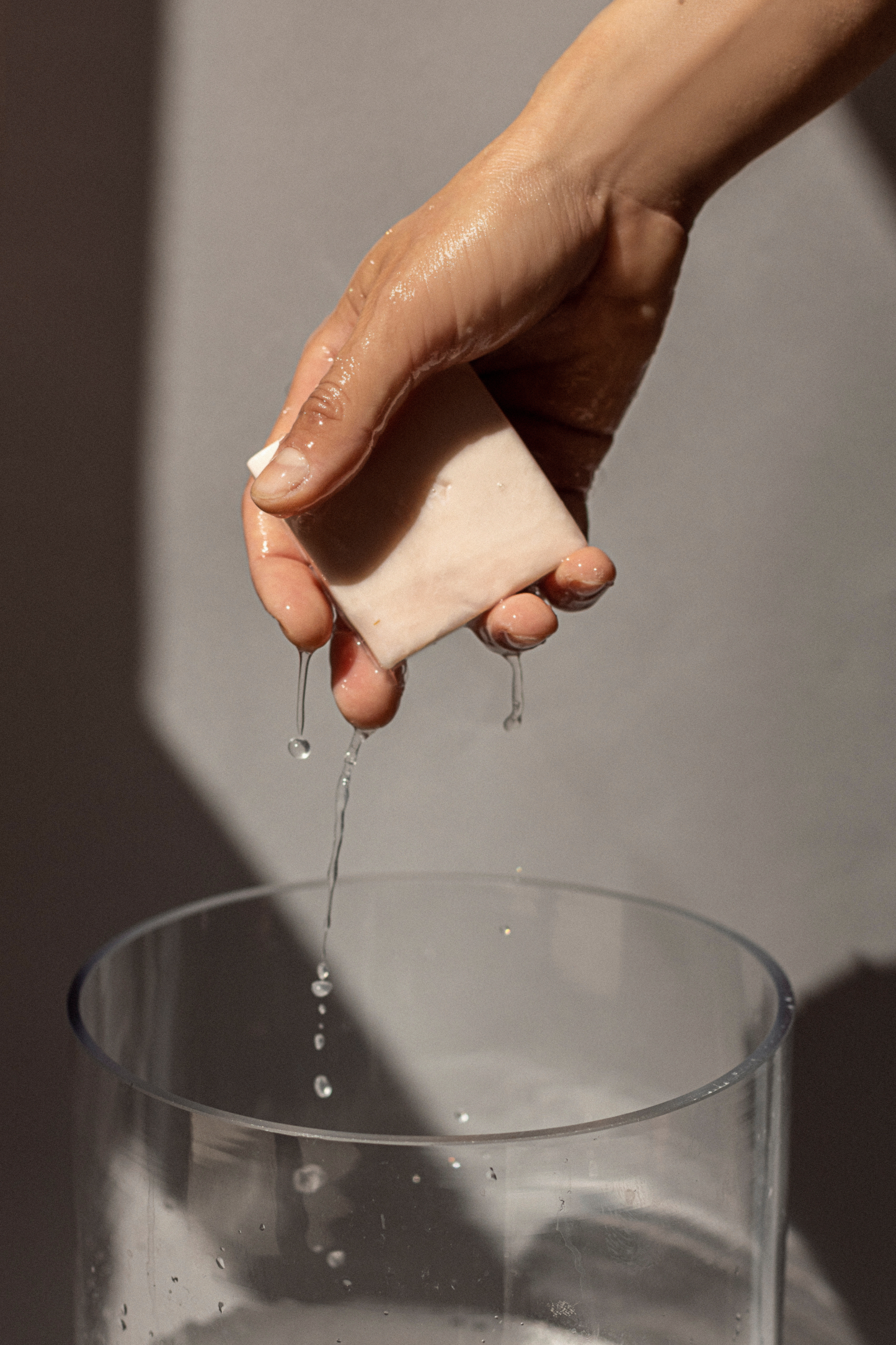 Woman's wet hand holding Ondo Beauty 36.5's Calamind & Oatmeal Soothing Cleansing Bar with water dripping down, against a grey background.