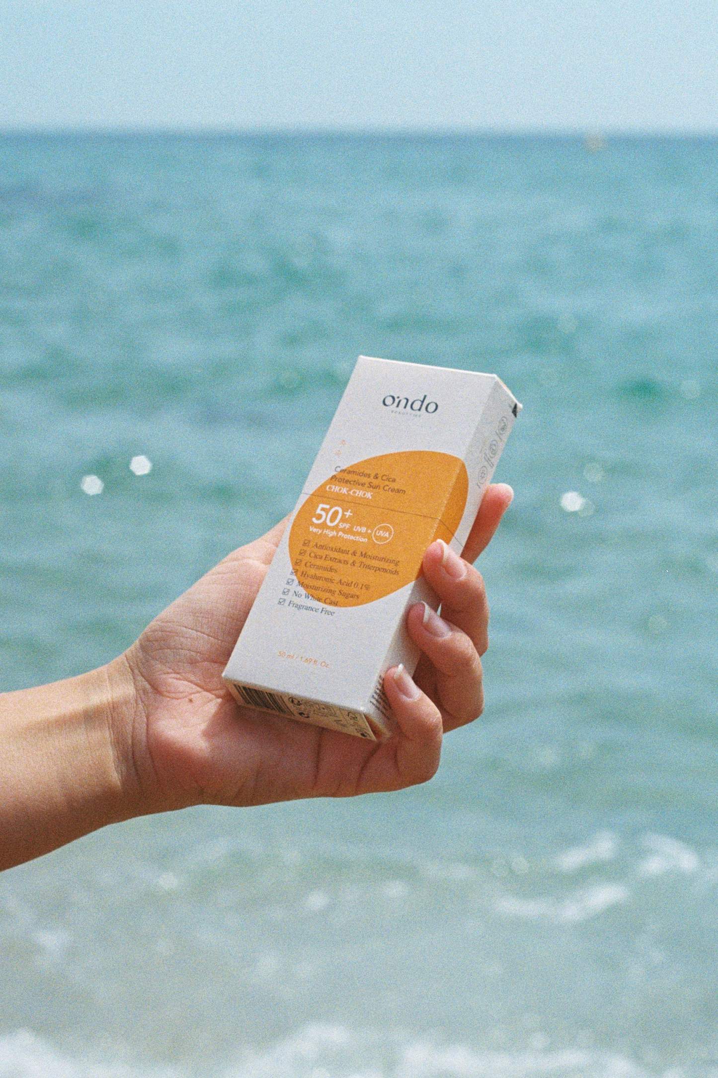Hand holding a box containing Ondo Beauty 36.5's Ceramides & Cica Protective Sun Cream with the sea in the background. Available at Sinless Skincare.