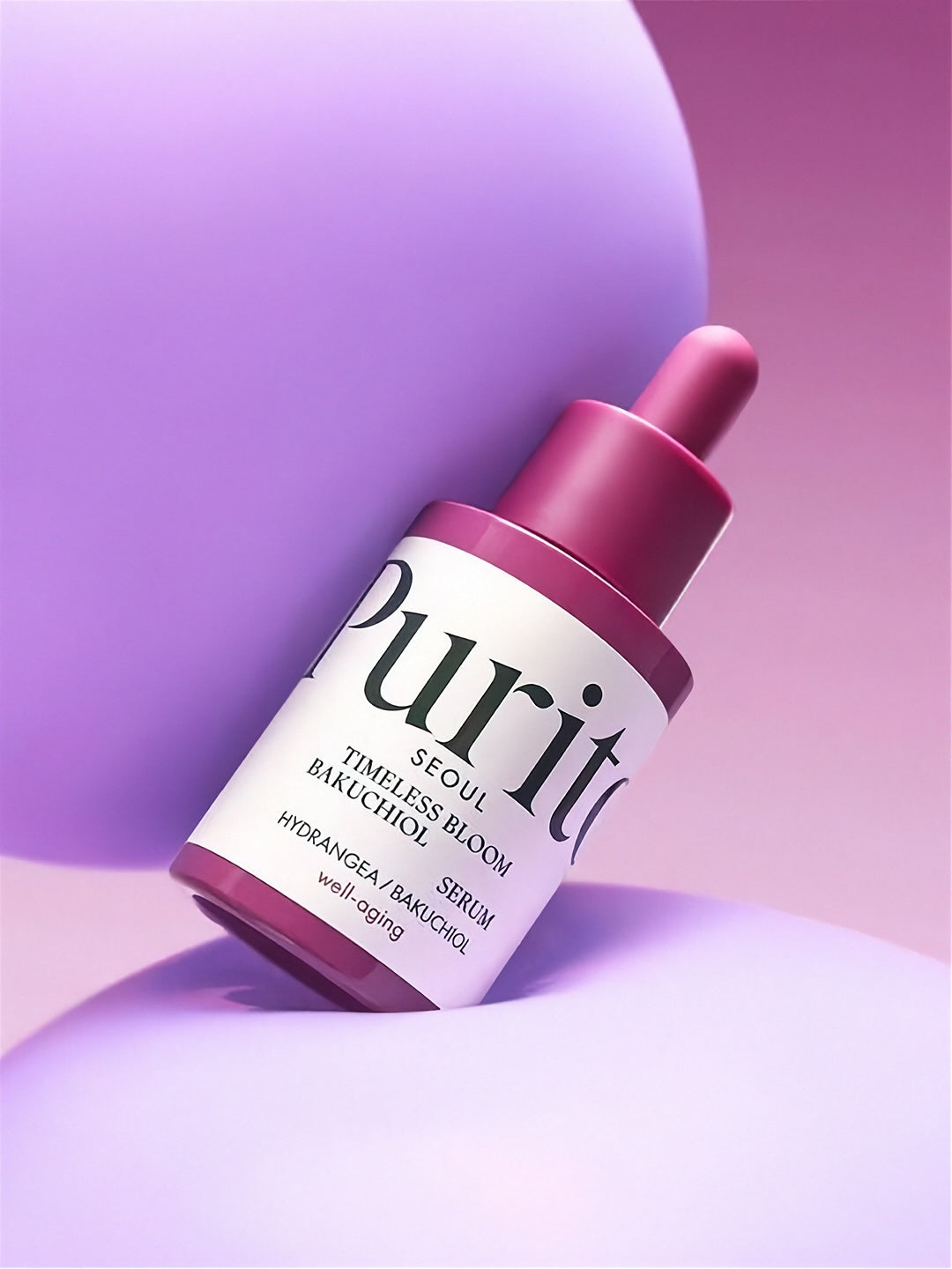 Bottle of Purito Timeless Bloom Backuchiol Serum between two purple balloons, against a pink background