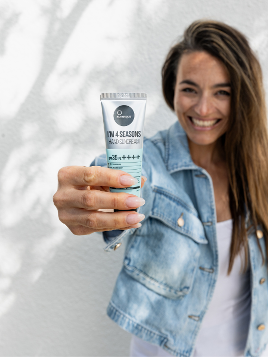Woman in denim jacket holding a tube of Suntique's I'm 4 Seasons Hand Sun Cream in her outstretched hand, against a white background. Available at Sinless Skincare.