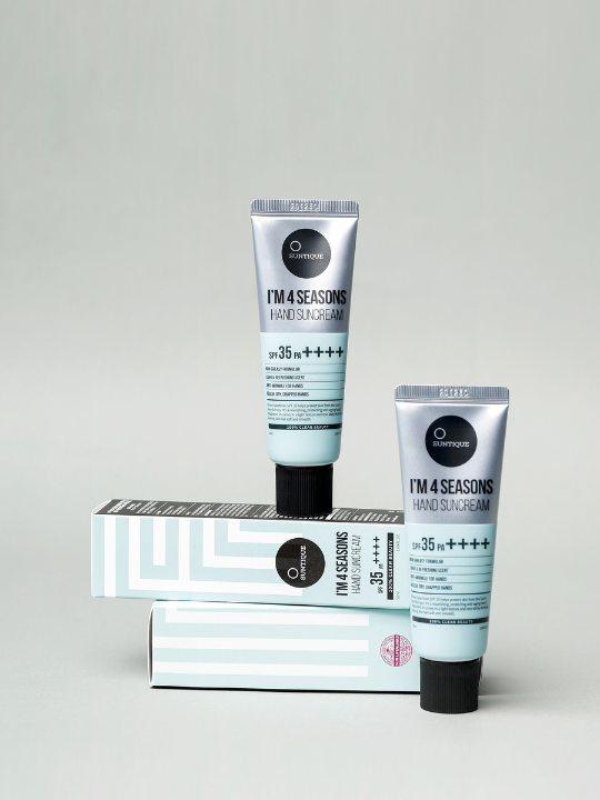 Two tubes of Suntique's I'm 4 Seasons Hand Sun Cream standing upright, their boxes lying sideways, against a light grey background. Available at Sinless Skincare.