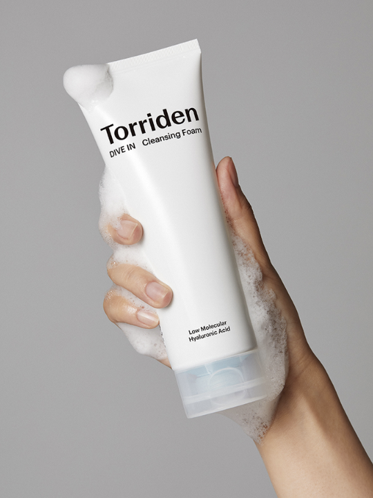 Hand holding a tube of Torriden Dive In Cleansing Foam, with foaming bubbles around the hand and bottle, against a grey background. Available at Sinless Skincare.