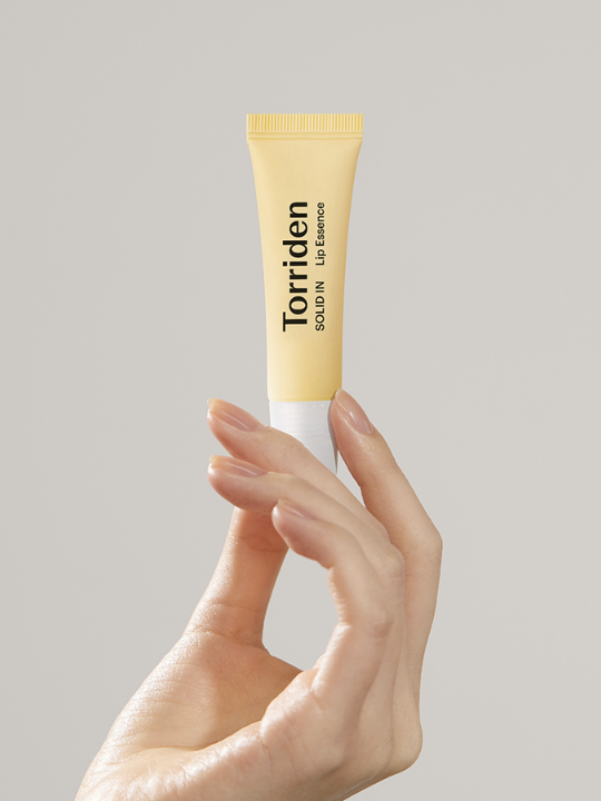 Woman's hand holding up a tube of Torriden Solid In Lip Essence, against a light grey background. Available at Sinless Skincare.