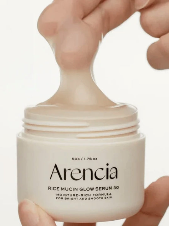 A close-up of a hand holding an open jar of Arencia Rice Mucin Glow Serum 30, with a thick beige gel being lifted from the jar against a clean white background.