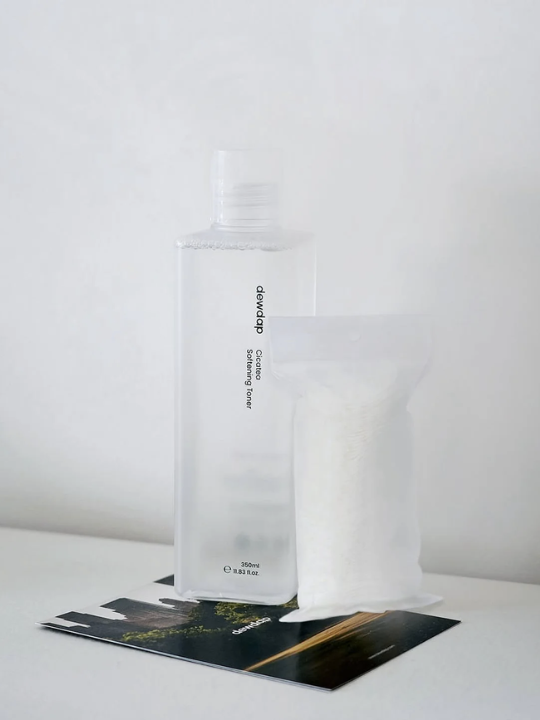 Bottle of Dewdap Cicatea Sofening Toner standing on a brand flyer on a table, with a clear refill bag next to it.
