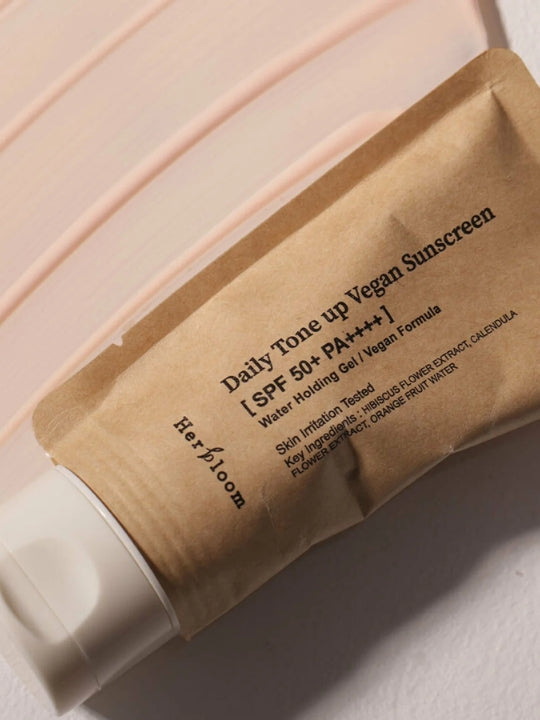 A squeeze tube of Herbloom Daily Tone Up Vegan Sunscreen SPF 50+ PA+++, shown diagonally on a light pink creamy surface.