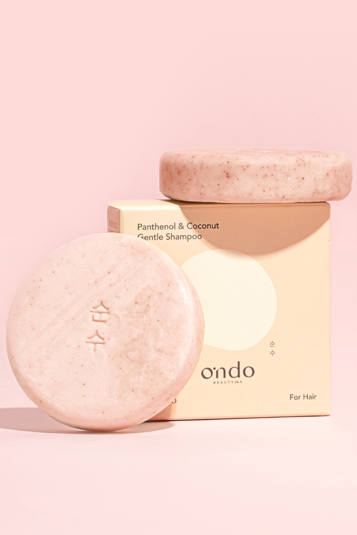 Close-up of two Ondo Beauty 36.5 Panthenol & Coconut Shampoo bars and their box on a pink background. One bar is resting on its side atop the upright box, while the other bar stands upright beside them. Available for purchase at Sinless Skincare.