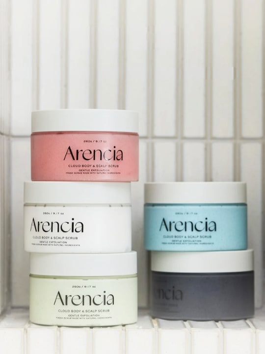 Five jars of Arencia Cloud body and scalp scrubs stacked on a white tiled surface. The set includes a pink jar, a white jar, a light blue jar, a light green jar, and a dark gray jar, all with white lids.