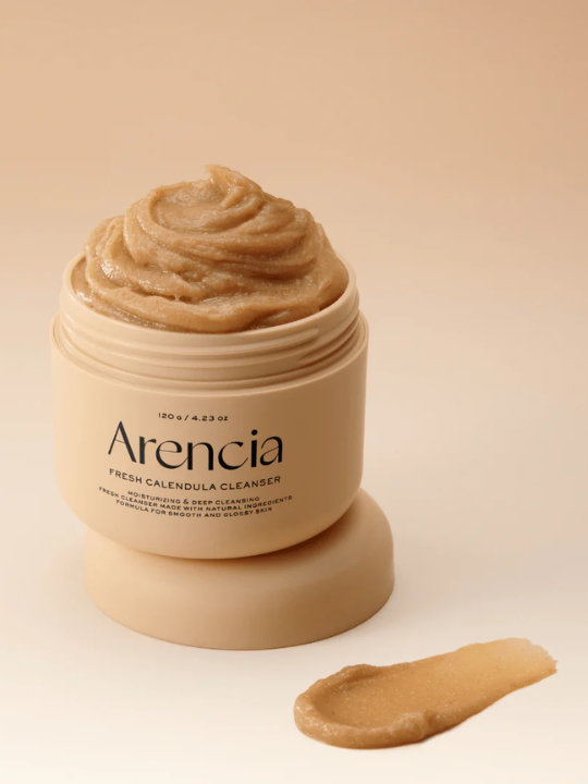 Open jar of Arencia Calendula rice mochi cleanser on beige background with swatch of product in front.
