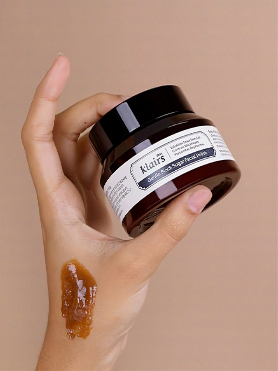 Hand holding a jar of Klairs Gentle Black Facial Polish with a swatch on the hand against a beige background