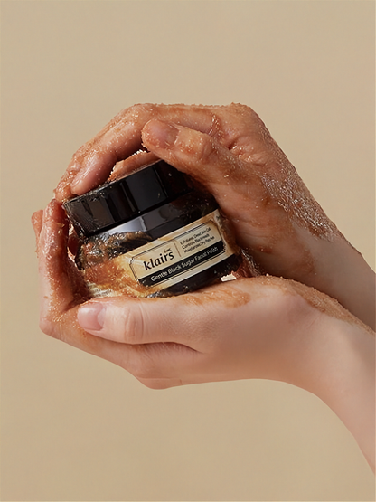Hands holding a jar of Klairs Gentle Black Facial Polish with brown substance covering the hands, against a beige background.