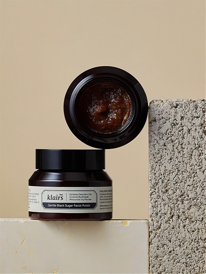 Two Klairs Gentle Black Facial Polish jars, one with open lid on a textured stone surface and beige background.