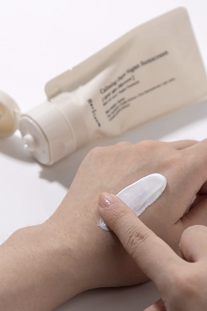 Herbloom's Calming Days Vegan Sunscreen is applied to a woman's hands close up. An open tube of the product lying on its side is blurred in the background.