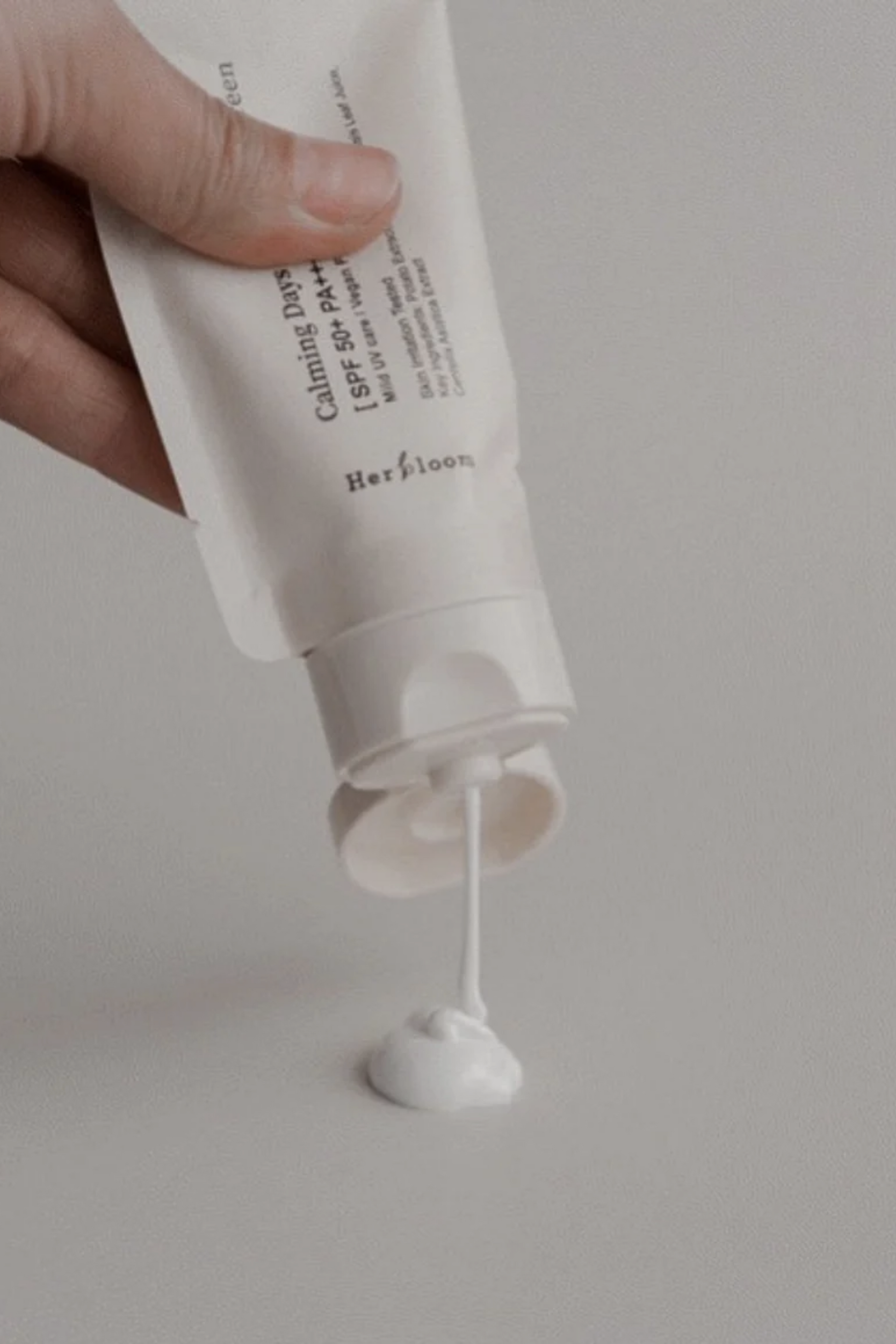An open bottle of Herbloom's Calming Days Vegan Sunscreen is being squeezed upside down onto a light grey surface.