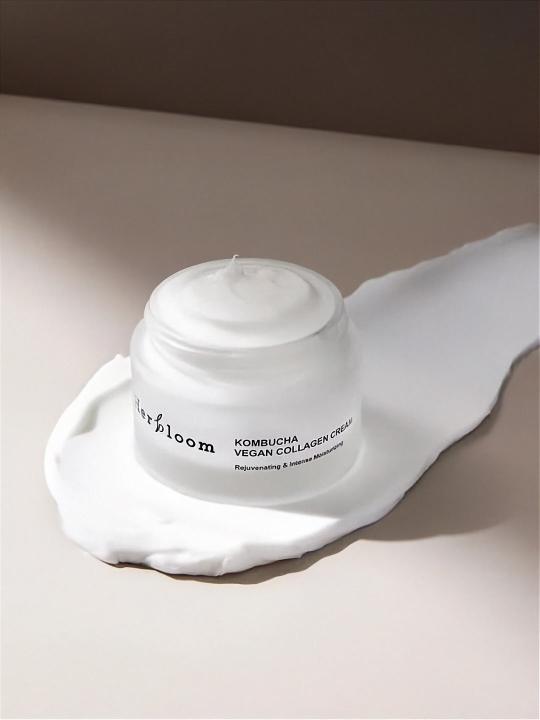 Open jar of Herbloom's Kombucha Vegan Collagen Cream on top of a large smear of white cream, on a neutral surface and background.
