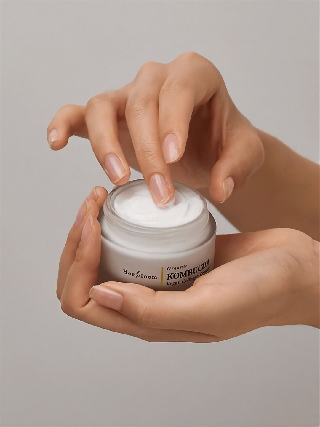 Fingers dipping into an open jar of Herbloom's Kombucha Vegan Collagen Cream against a light grey background