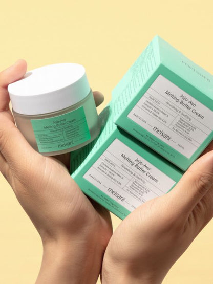 Two hands holding a jar of Meisani Jojo-Avo Melting Butter Cream and two of its retail boxes - on a yellow background