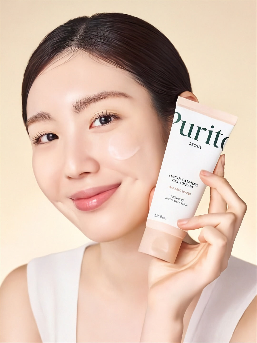 Woman holding up tube of Purito Seoul Oat In Calming Gel Cream against her cheek on a beige background