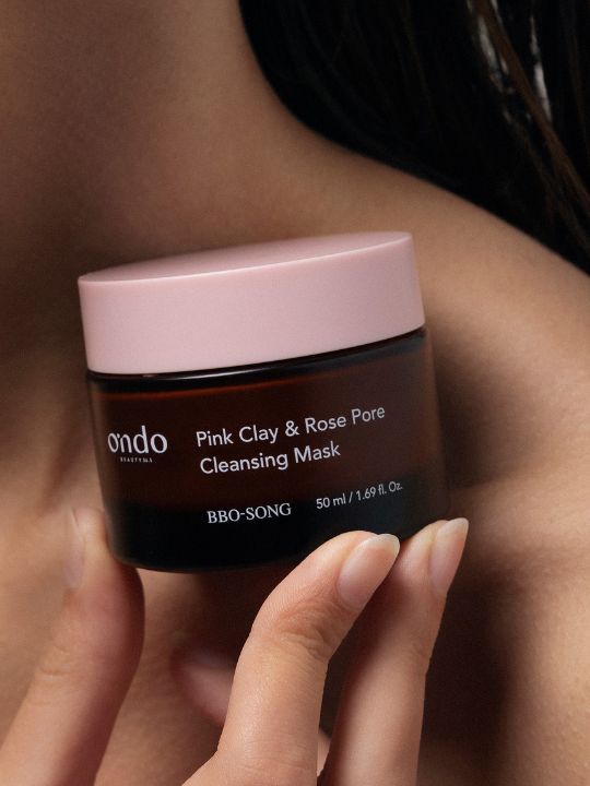 Hand holding a jar of Ondo Beauty Pink Clay & Rose Pore Cleansing Mask  against a bare shoulder