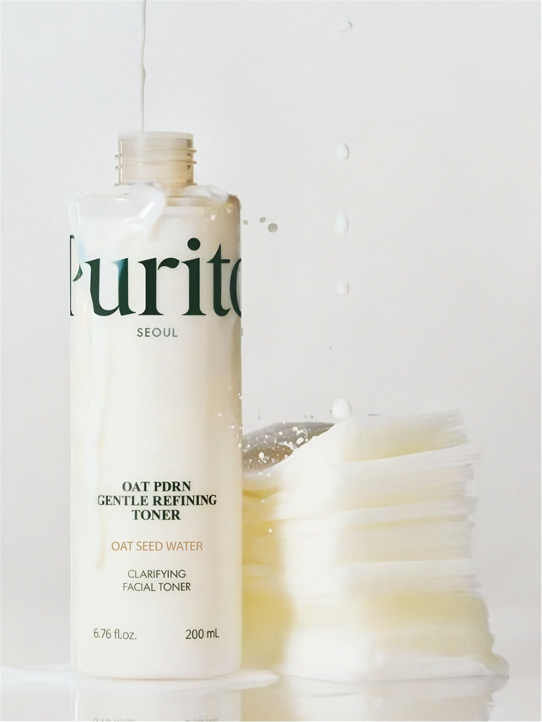 Purito Oat PDRN Gentle Refining Toner bottle next to cotton pads soaked in liquid on a white background