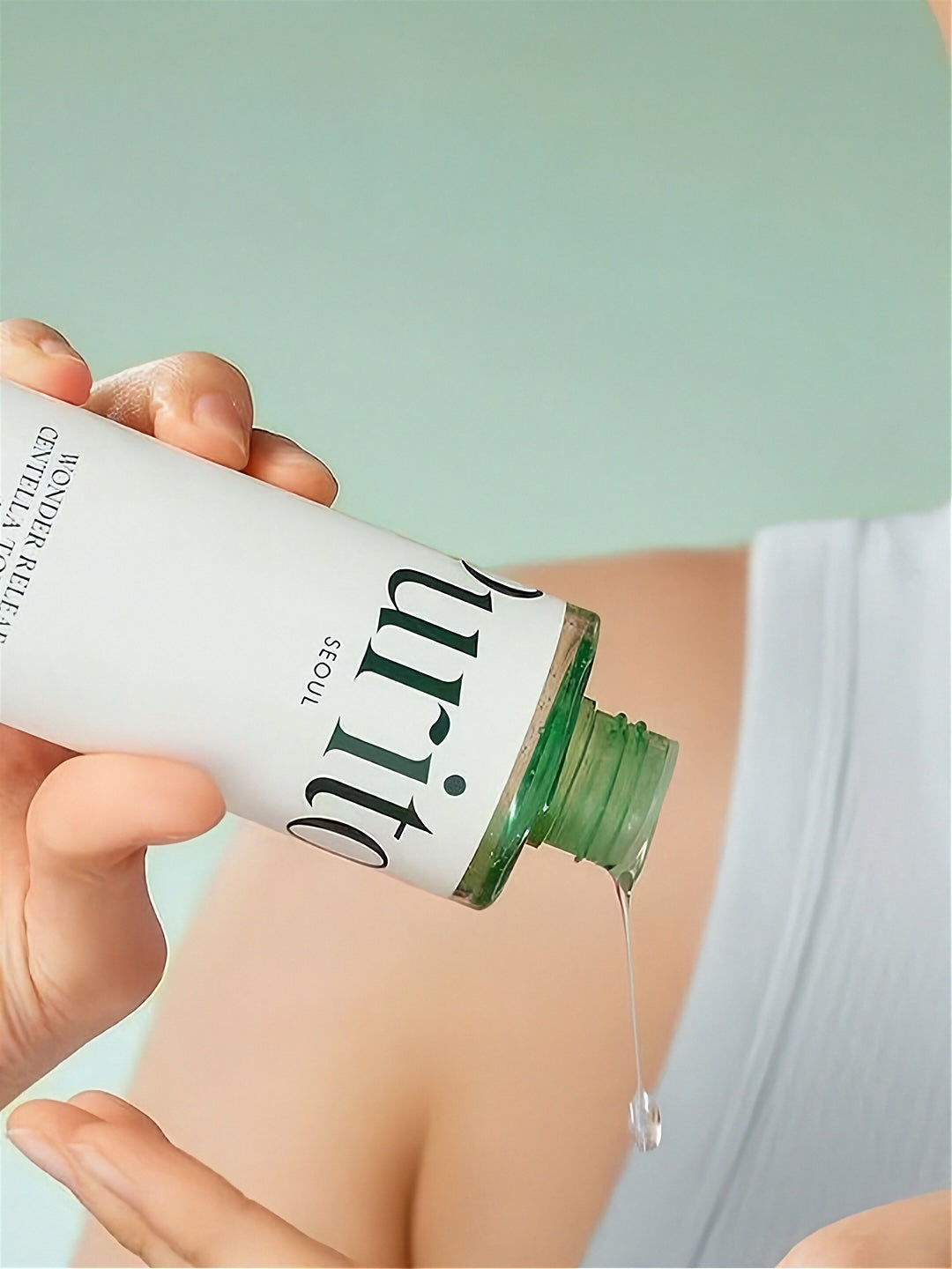 Hand pouring a bottle of Purito Wonder Releaf Centella Toner Unscented, against a light green background.