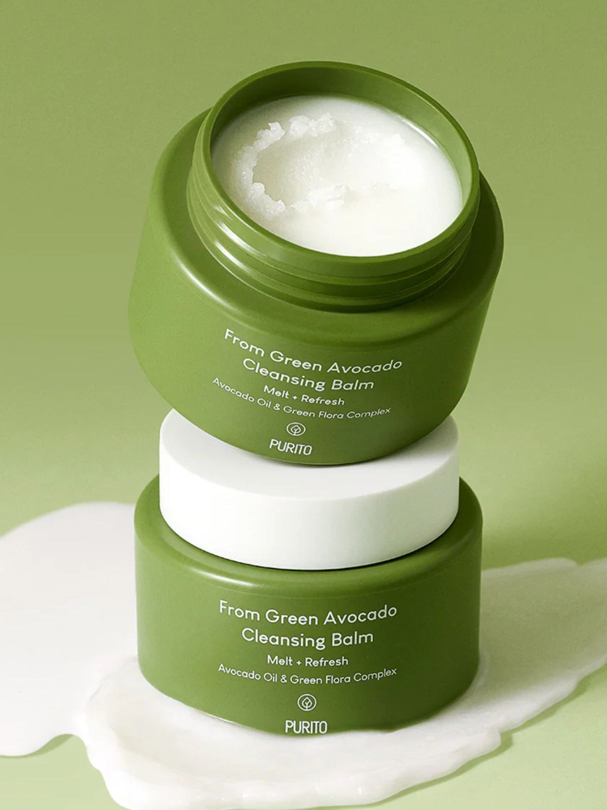 Two green PURITO jars of From Green Avocado Cleansing Balm stacked on a light green background, with one jar open to show the white balm inside and a white cream spill in front.
