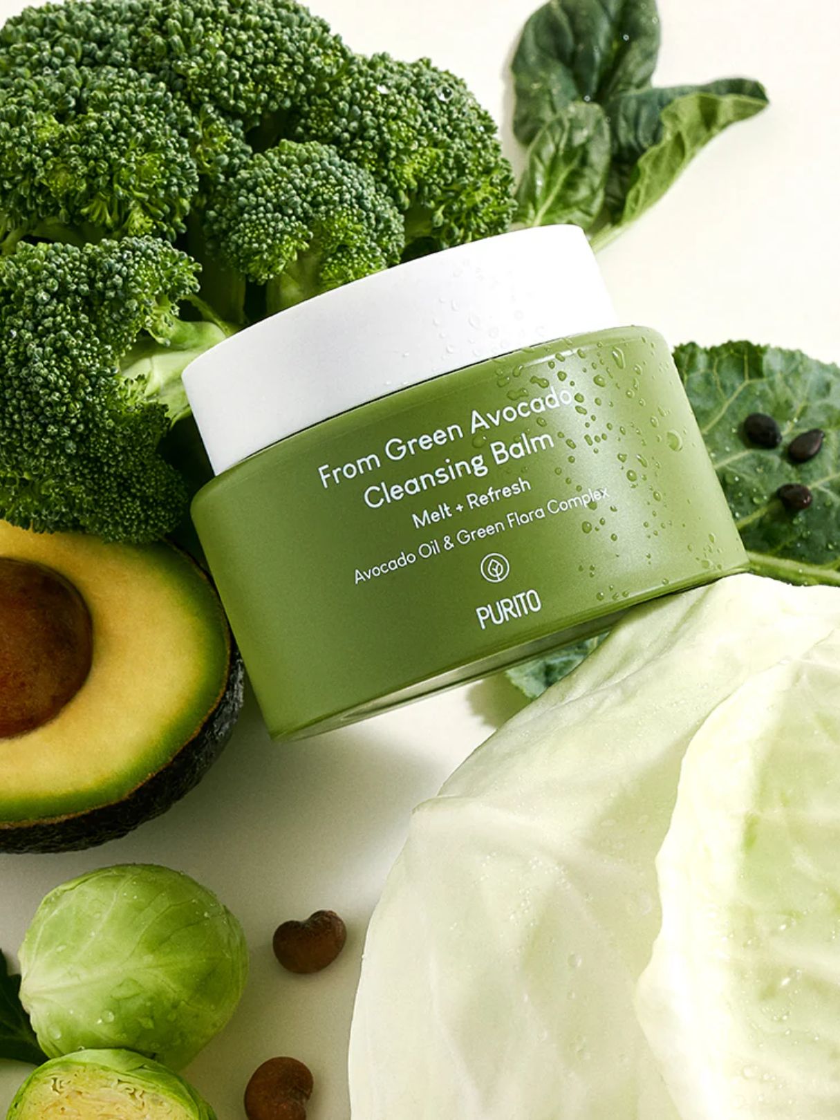 Purito From Green Avocado Cleansing Balm in a green jar with a white lid, styled with fresh broccoli, avocado, cabbage, and Brussels sprouts on a white background.