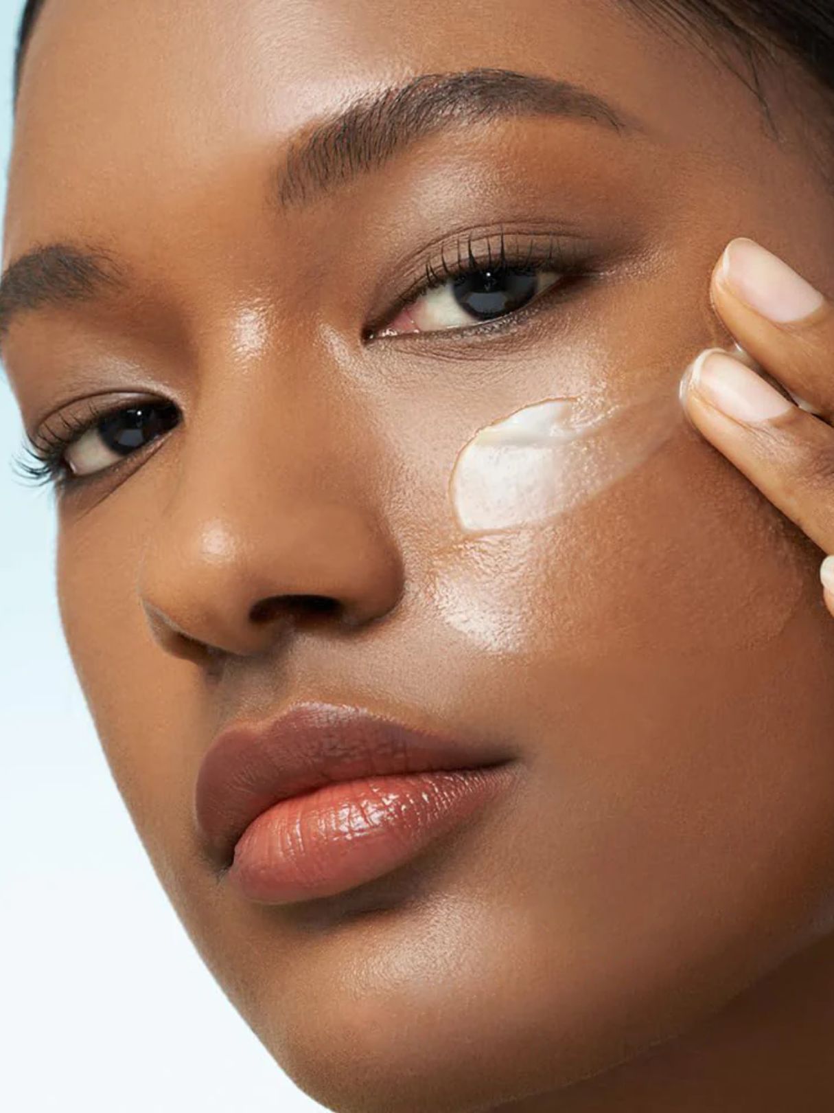 Close-up of a woman applying Purito From Green Avocado Cleansing Balm to her cheek, showing a smooth, glossy skin texture against a light blue background.