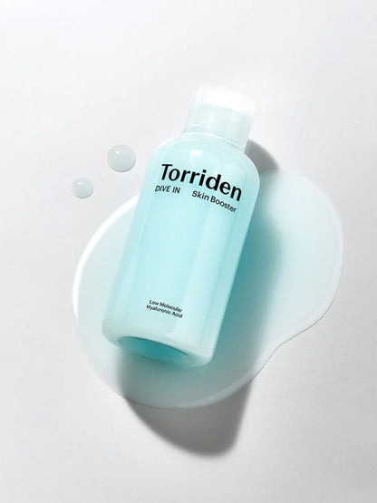A bottle of Torriden Dive In Skin Booster in a pale aqua blue color, styled on a light grey surface with a white liquid splash and small droplets.