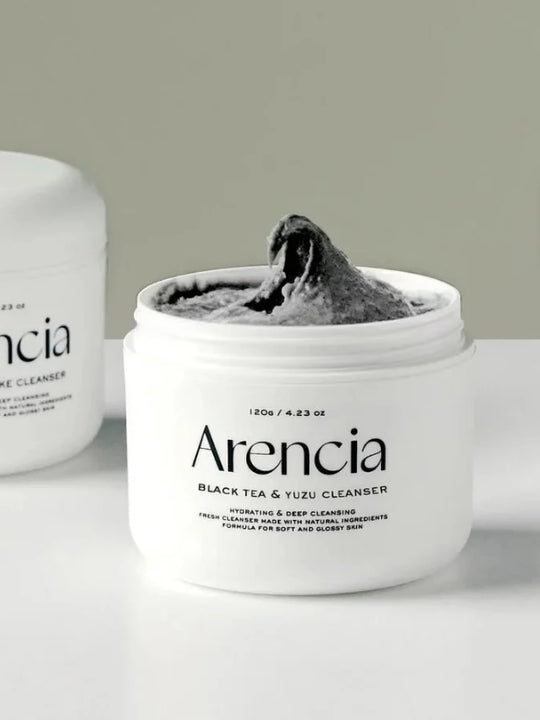 An open white jar of Arencia Black Tea & Yuzu Cleanser showing a thick dark gray cleanser inside, with a second blurred jar in the background on a light surface against a pale gray wall.