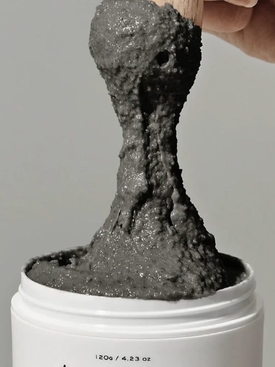 A close-up of a thick, dark gray slime-like substance being lifted from a white jar by a hand using a wooden stick, against a plain light gray background.
