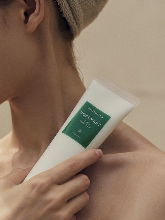 Woman holding a tube of Aromatica Rosemary 3 in 1 Treatment to her neck against a neutral background
