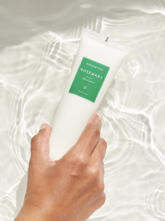 Hand holding a bottle of Aromatica Rosemary 3 in 1 Treatment against a white, watery background