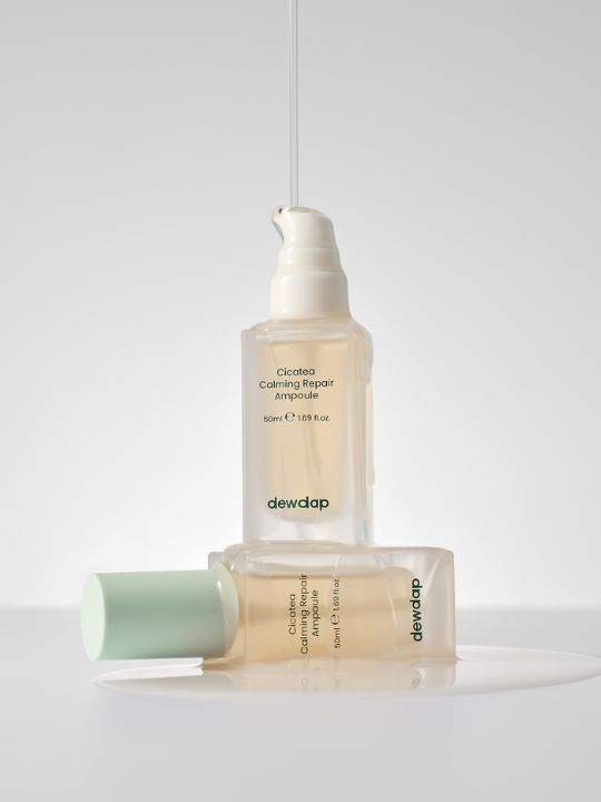 Two bottles of Dewdap Cicatea Calming Repair Ampoule on a grey background with liquid pouring down