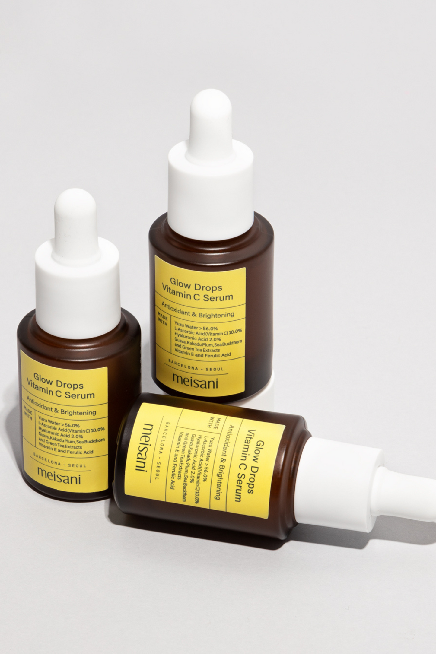 Three bottles of Meisani's Glow Drops Vitamin C Serum against a light grey background. One bottle is lying on its side. Available for purchase at Sinless Skincare.