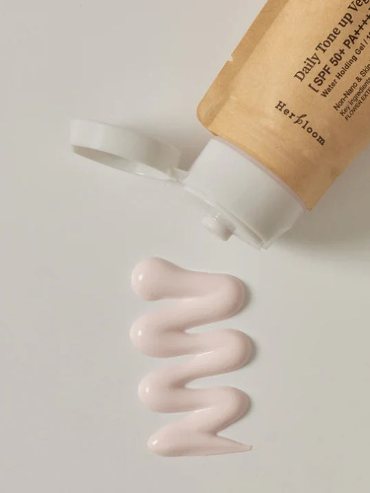 A squeeze tube of Herbloom Daily Tone Up Vegan Sunscreen SPF 50+ lies on its side with the cap off, showing a wavy squiggle of pale pink cream below it on a light gray background.