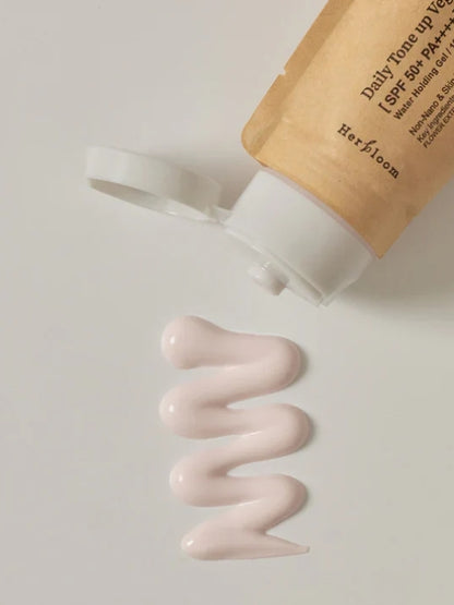 A squeeze tube of Herbloom Daily Tone Up Vegan Sunscreen SPF 50+ lies on its side with the cap off, showing a wavy squiggle of pale pink cream below it on a light gray background.