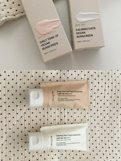 Two Herbloom sunscreens shown in a split view: boxed Daily Tone Up Vegan Sunscreen and Calming Days Vegan Sunscreen, plus the corresponding squeeze tubes, arranged on a light surface with a textured fabric backdrop.