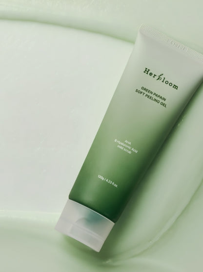 A squeeze tube of Herbfloom Green Papain Soft Peeling Gel resting diagonally on a smooth, light green surface with a matching liquid gel visible in the background.