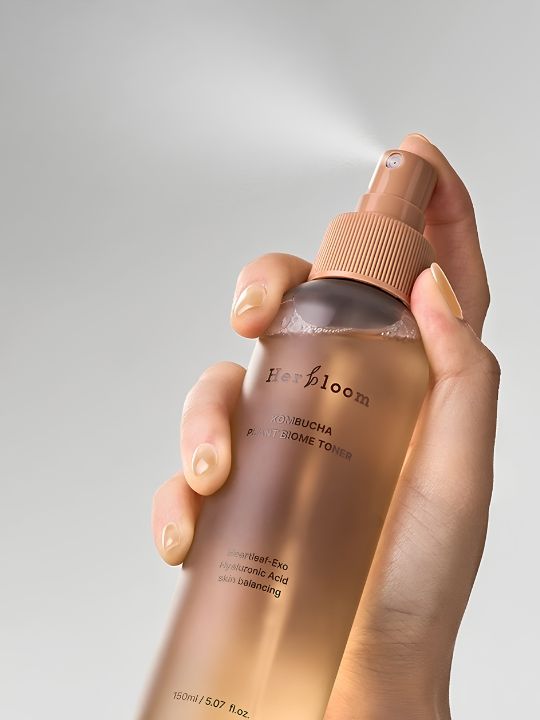 Close up of a hand spraying a bottle of Herbloom Kombucha Plant Biome Toner against a grey background