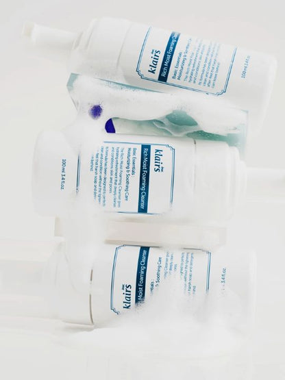 Three bottles of Klairs Rich Moist Foaming Cleanser stacked on their sides with white foam surrounding them on a white background