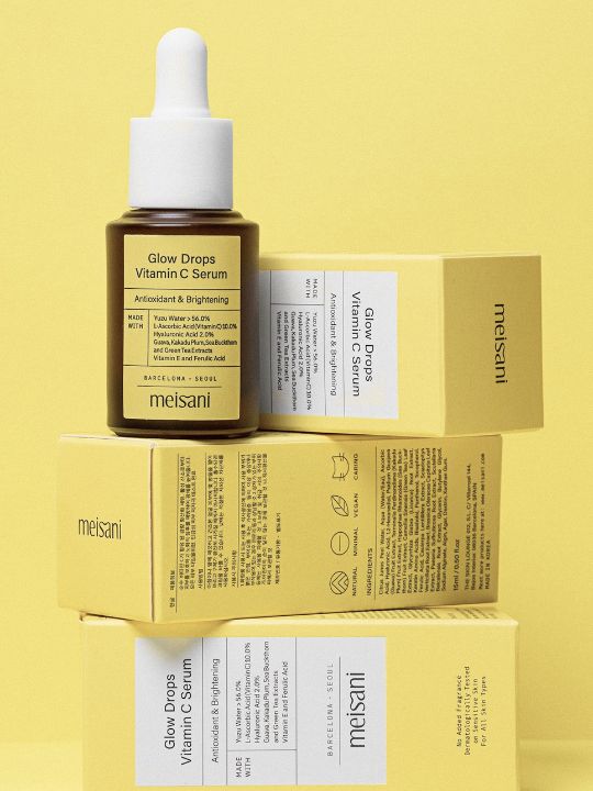 Meisani Glow Drops Vitamin C Serum standing on its retail boxes against a yellow background