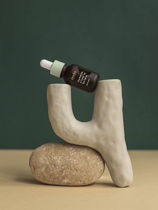 Ondo Beauty Niacinamide & Green Tea Eye Serum dropper bottle balancing on a stone against a green background