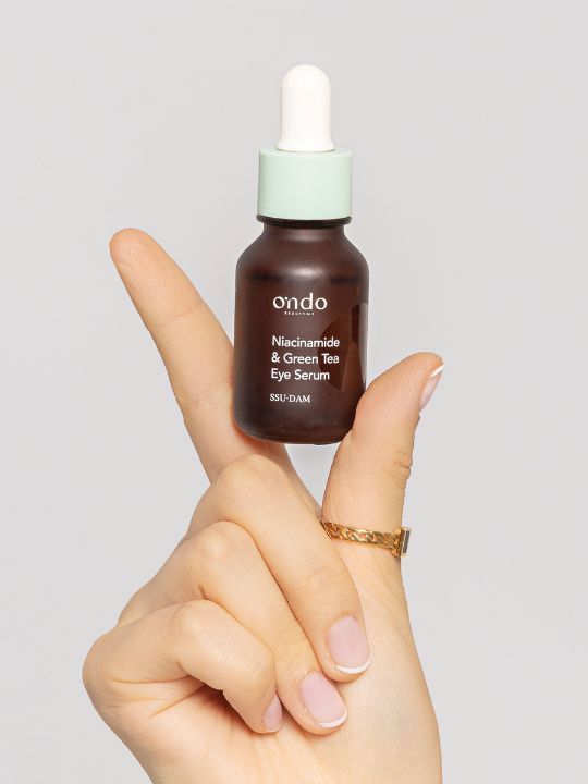 Hand holding up a bottle of Ondo Beauty Niacinamide & Green Tea Eye Serum on a light grey background
