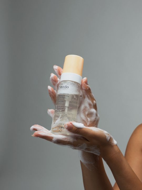 Person with foamy hands holding a bottle of Ondo Beauty 36.5 PHA & Saponaria Purifying Foaming Cleanser against a grey background