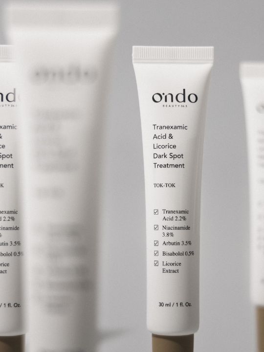 Ondo Beauty Tranexamic Acid & Licorice Dark Spot Treatment tube with blurred tubes on a grey background