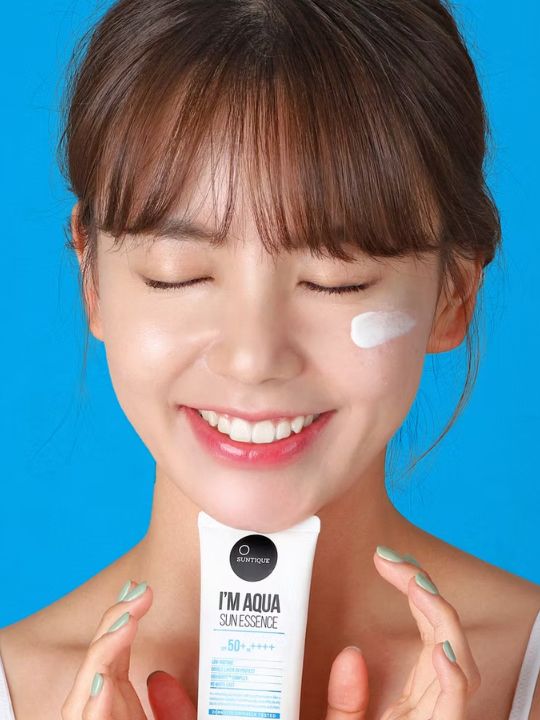 Woman holding tube of Suntique I'm Aqua Sun Essence with a swatch on her cheek, against a blue background